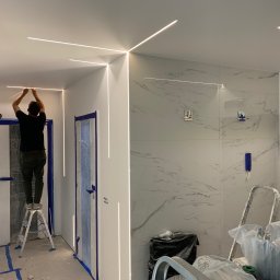 Piotr Warchoł - Instalacja liniowego oświetlenia LED na suficie w pomieszczeniu w trakcie remontu, widoczny mężczyzna na drabinie, zabezpieczone folią drzwi i marmurowe płytki na ścianie.