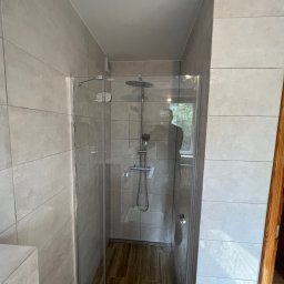 Piotr Warchoł - Nowoczesna kabina prysznicowa z deszczownicą i szklanymi drzwiami, wykończona płytkami w odcieniach szarości, z drewnopodobną podłogą i odbiciem sylwetki fotografa w szkle.