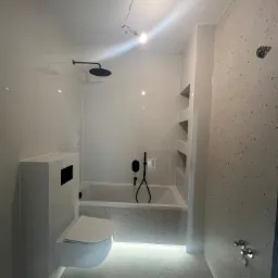 Nowoczesna łazienka z białymi błyszczącymi płytkami i czarną armaturą, podświetlana wanna i toaleta podwieszana, ściana z mozaiką.