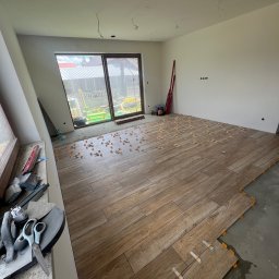 Piotr Warchoł - Układanie podłogi z płytek imitujących drewno, widoczne krzyżyki dystansowe, narzędzia glazurnicze na parapecie, surowy stan ścian i podciągnięte kable.