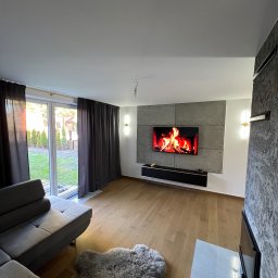 Piotr Warchoł - Nowoczesny salon z drewnianą podłogą, szarą sofą narożną, dywanem z długim włosiem i ścianą telewizyjną z imitacją betonu, podświetloną kinkietami.