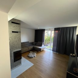 Piotr Warchoł - Jasne wnętrze salonu z drewnianą podłogą, szarą sofą narożną, kominkiem obłożonym szarym kamieniem dekoracyjnym i dużym oknem balkonowym z zasłonami w kolorze grafitowym.