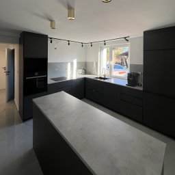 Piotr Warchoł - Nowoczesna, minimalistyczna kuchnia z czarnymi szafkami i szarą wyspą, oświetlona złotymi lampami sufitowymi i światłem dziennym wpadającym przez okno.