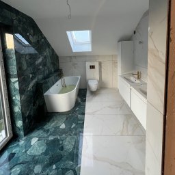Piotr Warchoł - Nowoczesna łazienka na poddaszu z oknem dachowym, wykończona płytkami w dwóch kolorach: zielonym marmurze i białym marmurze z beżowymi żyłami, z wanną wypełnioną wodą, toaletą i podwójną umywalką...