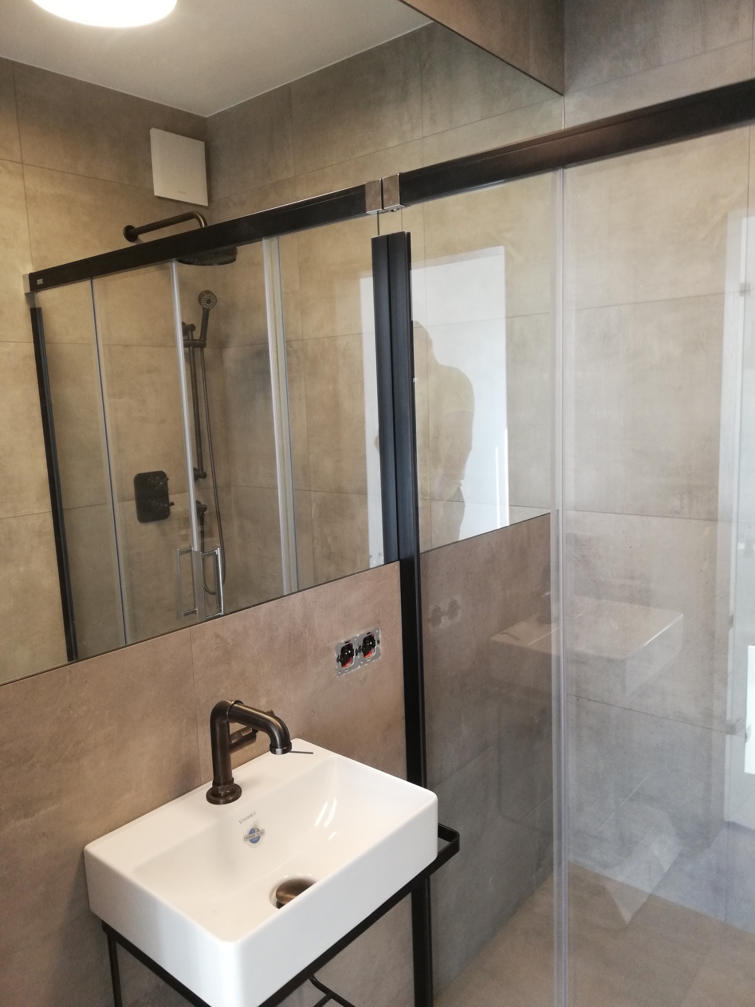 Nowoczesna łazienka z kabiną prysznicową typu walk-in, umywalką nablatową na metalowym stelażu i szarą ceramiką ścienną, widoczne niedokończone gniazdka elektryczne.