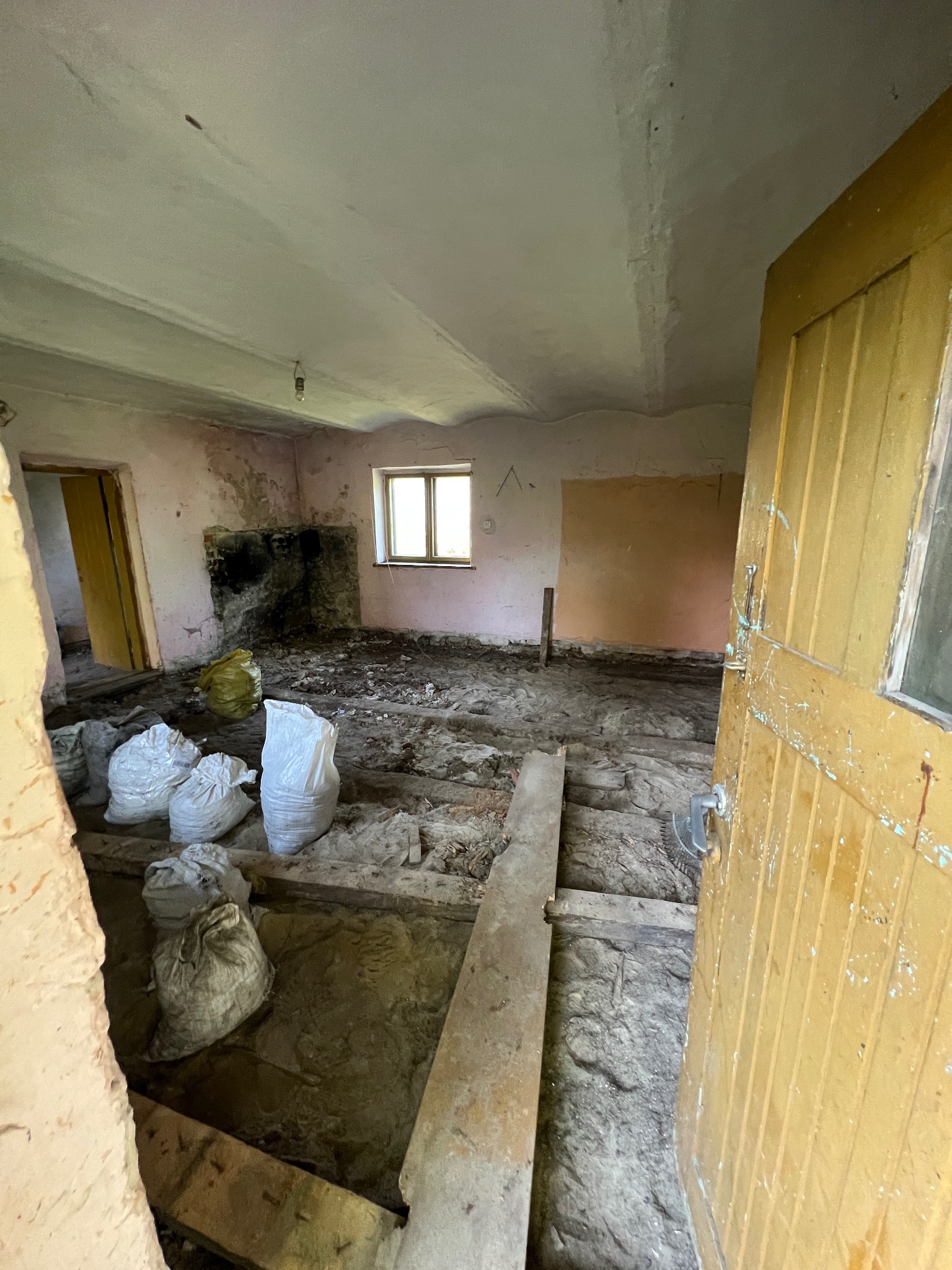 Pokój w trakcie remontu: zerwana podłoga, widoczne legary, worki z gruzem, ściany z resztkami farby i odsłoniętym murem, stare, żółte drzwi z widocznymi śladami użytkowania.