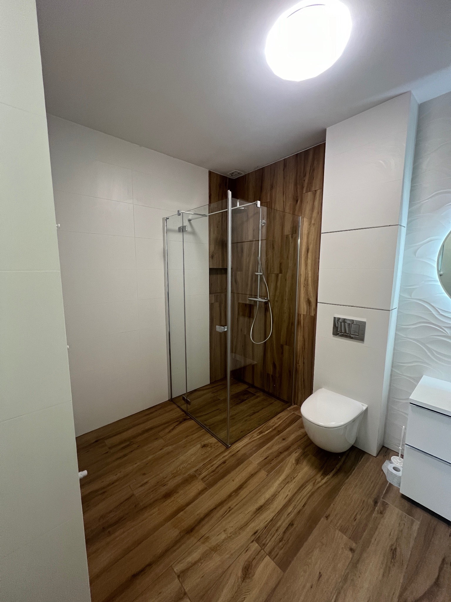 Nowoczesna łazienka z kabiną prysznicową typu walk-in, drewnopodobną podłogą, białą ceramiką i dekoracyjną ścianą z falistą strukturą.