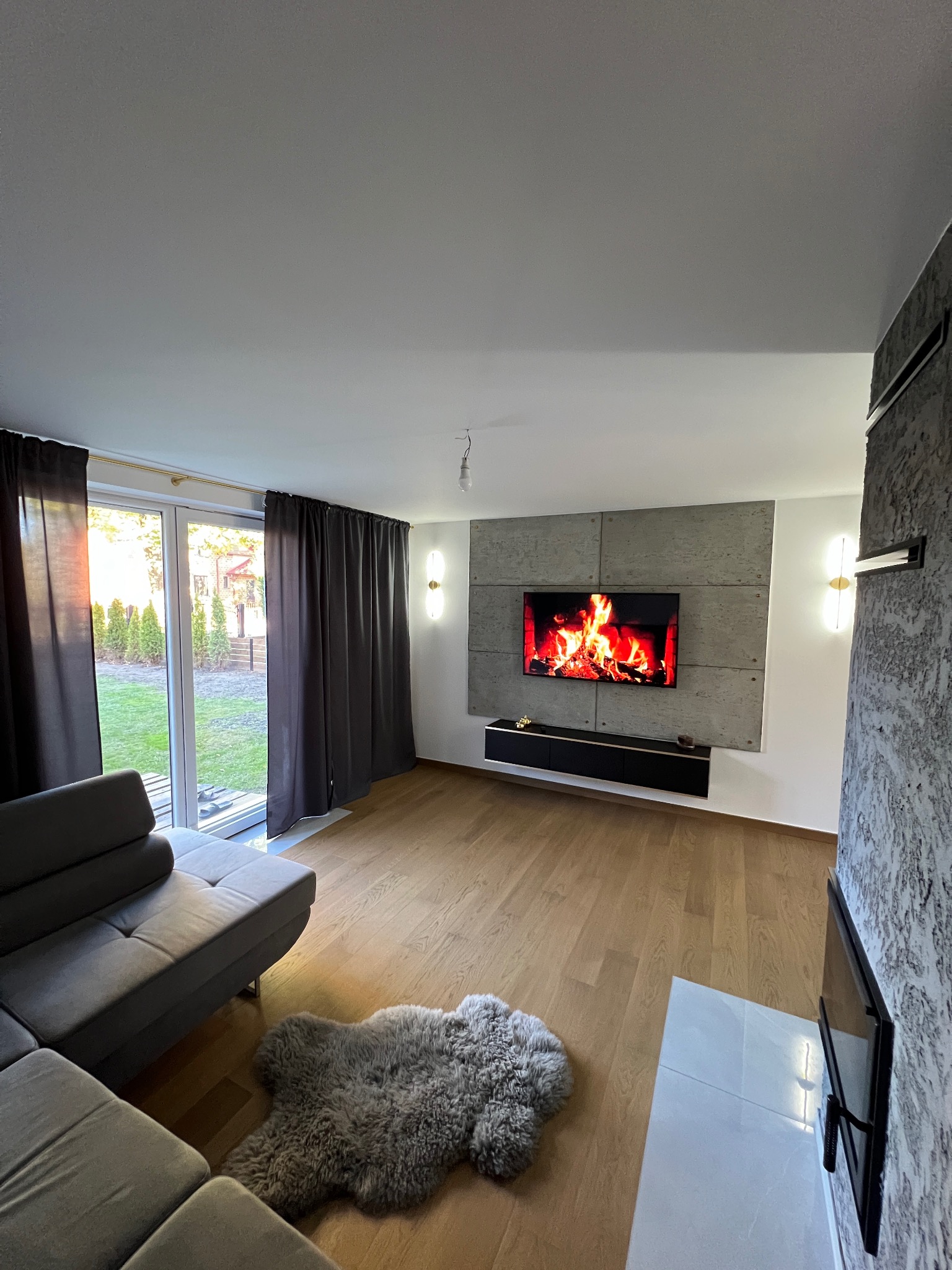 Nowoczesny salon z drewnianą podłogą, szarą sofą narożną, dywanem z długim włosiem i ścianą telewizyjną z imitacją betonu, podświetloną kinkietami.