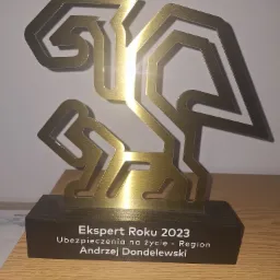 Złota statuetka 'Ekspert Roku 2023 Ubezpieczenia na życie - Region Andrzej Dondelewski' na drewnianej podstawie, stojąca na drewnianym biurku.