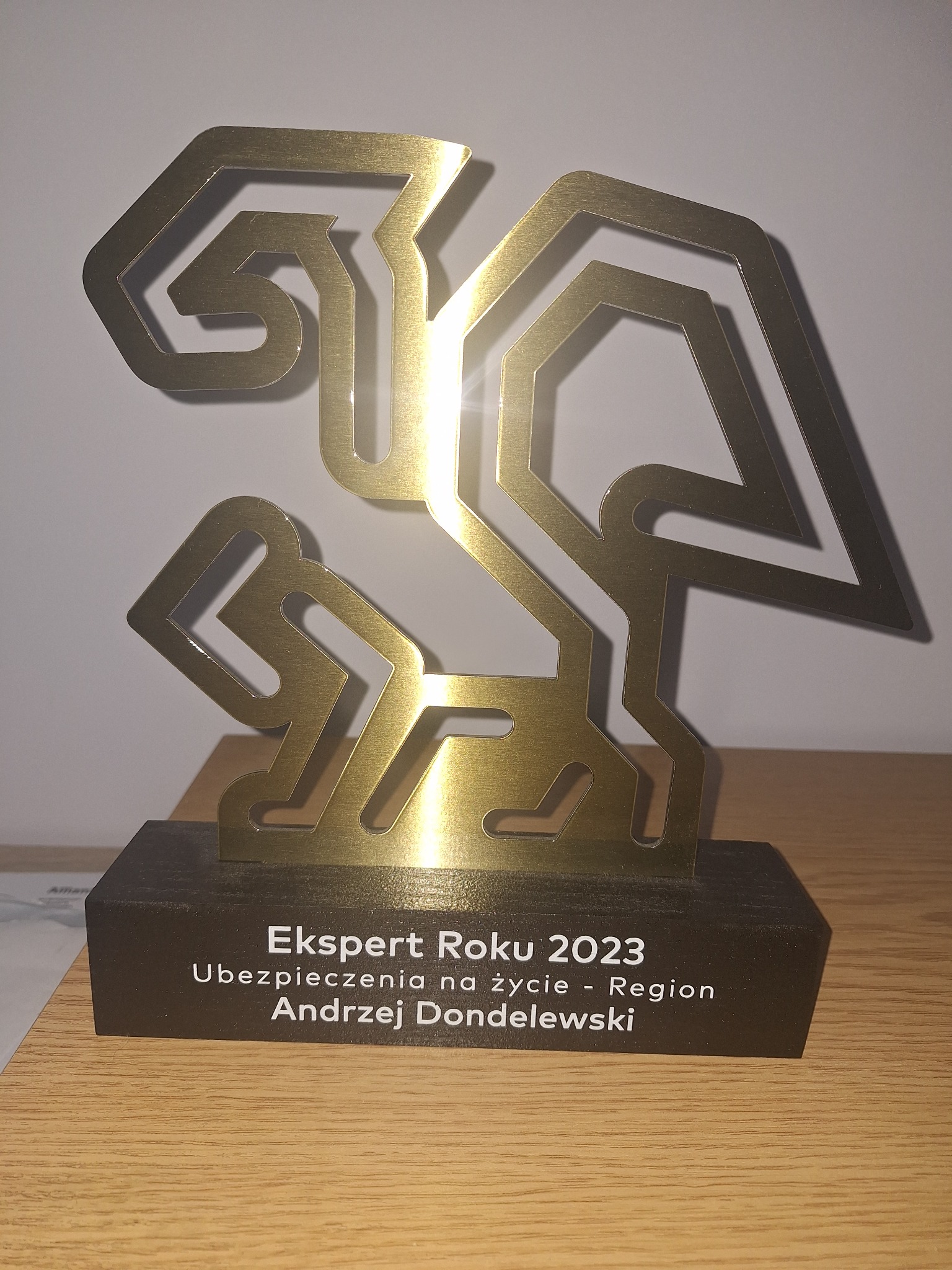 Złota statuetka 'Ekspert Roku 2023 Ubezpieczenia na życie - Region Andrzej Dondelewski' na drewnianej podstawie, stojąca na drewnianym biurku.