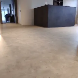 Jasnoszara, jednolita posadzka w nowoczesnym wnętrzu biurowym, widoczne minimalistyczne biurko recepcyjne w tle, ściany pomalowane na biało z subtelnym oświetleniem LED przy podłodze.