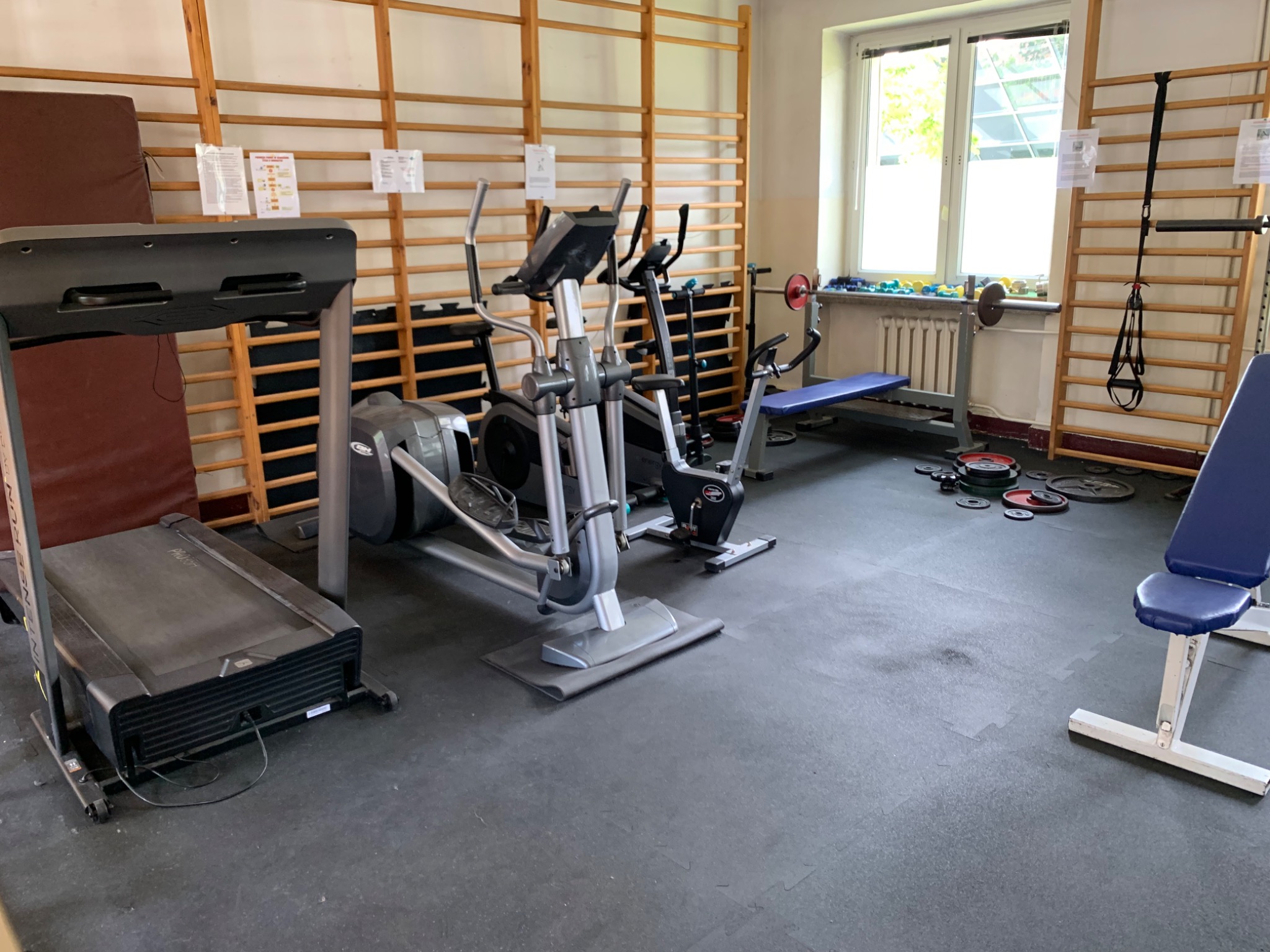 Ciemna, gumowa podłoga w siłowni z widocznymi śladami użytkowania, na której stoją różnorodne urządzenia do ćwiczeń: bieżnia, orbitrek, rowery stacjonarne oraz ławka do ćwiczeń z obciążeniami. Tło...