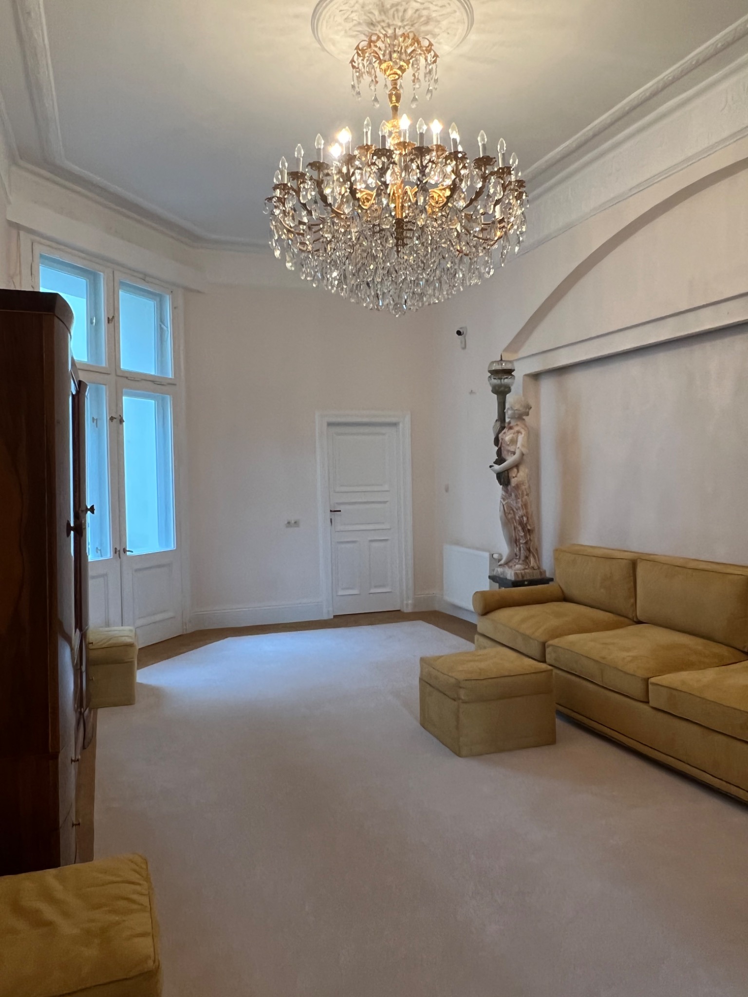 Jasnoszara wykładzina dywanowa w eleganckim salonie z kryształowym żyrandolem i sofą w odcieniach złota, widok na marmurową rzeźbę i drzwi balkonowe.