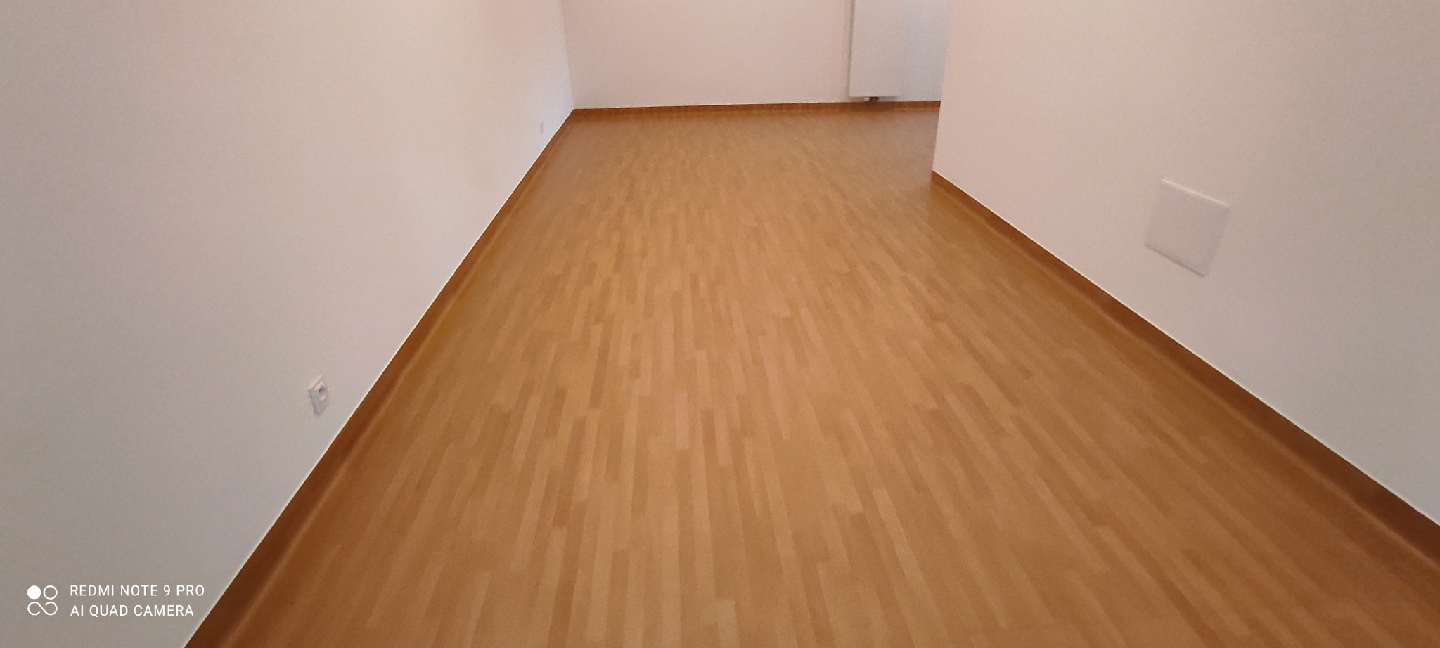 Jasna, drewnopodobna podłoga panelowa w pustym, świeżo wykończonym pokoju z białymi ścianami i gniazdkiem elektrycznym.