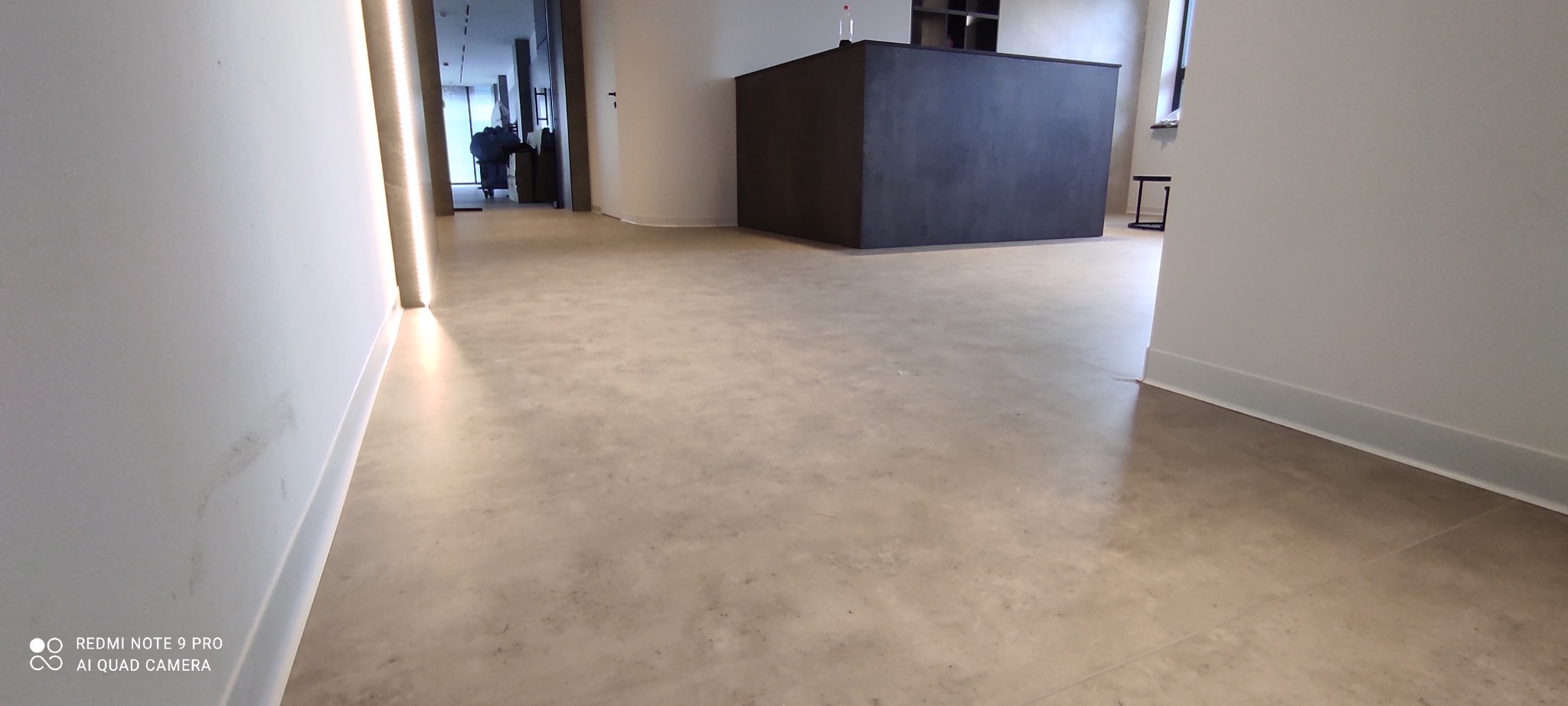 Jasnoszara, jednolita posadzka w nowoczesnym wnętrzu biurowym, widoczne minimalistyczne biurko recepcyjne w tle, ściany pomalowane na biało z subtelnym oświetleniem LED przy podłodze.