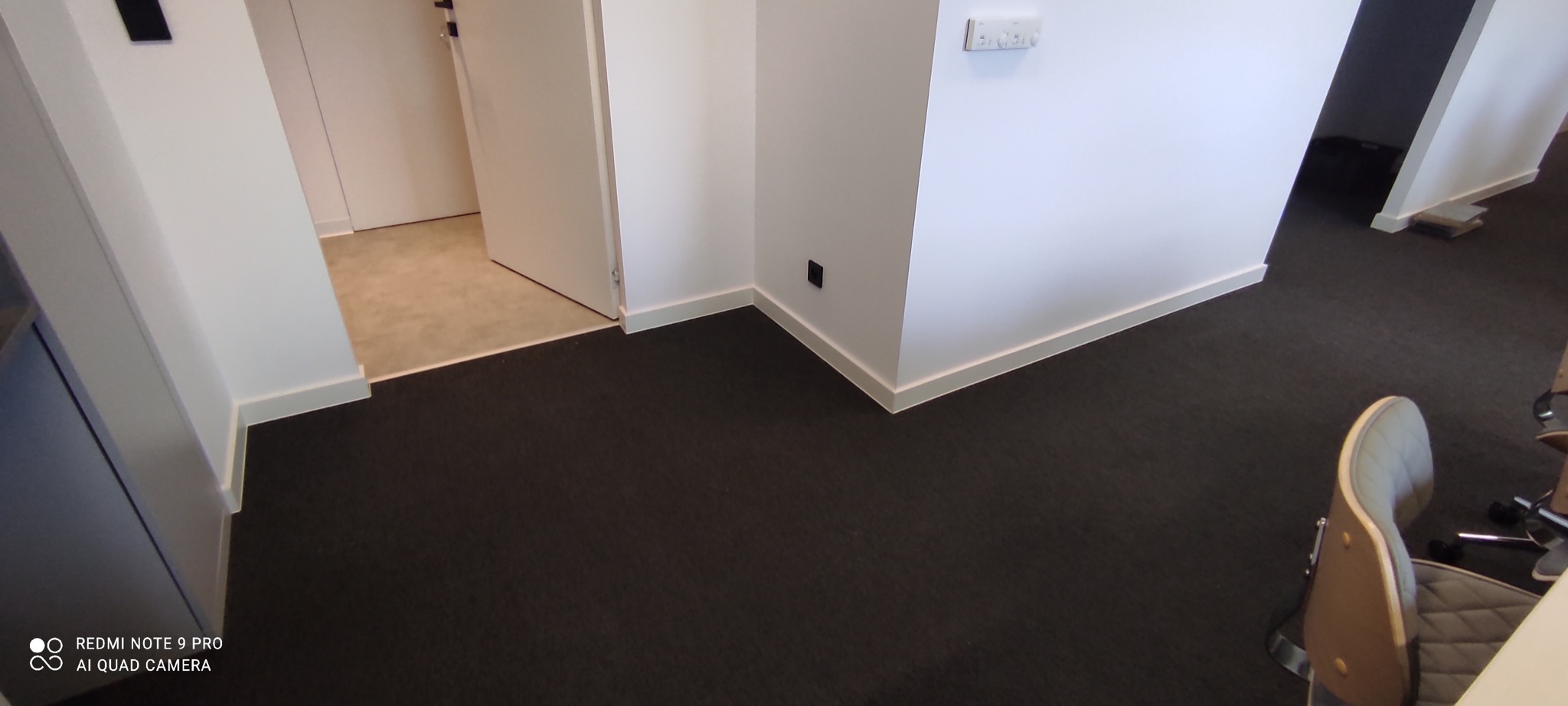 Ciemna wykładzina dywanowa w biurze z białymi ścianami i listwami przypodłogowymi, widoczny fragment krzesła biurowego.