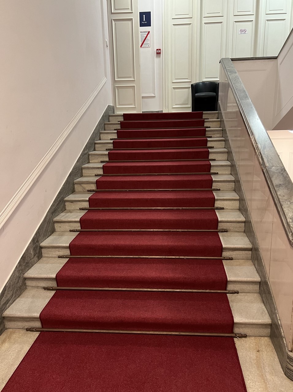 Czerwona wykładzina dywanowa zabezpieczona metalowymi prętami na marmurowych schodach w budynku z białymi panelami ściennymi i czarnym fotelem na górze.