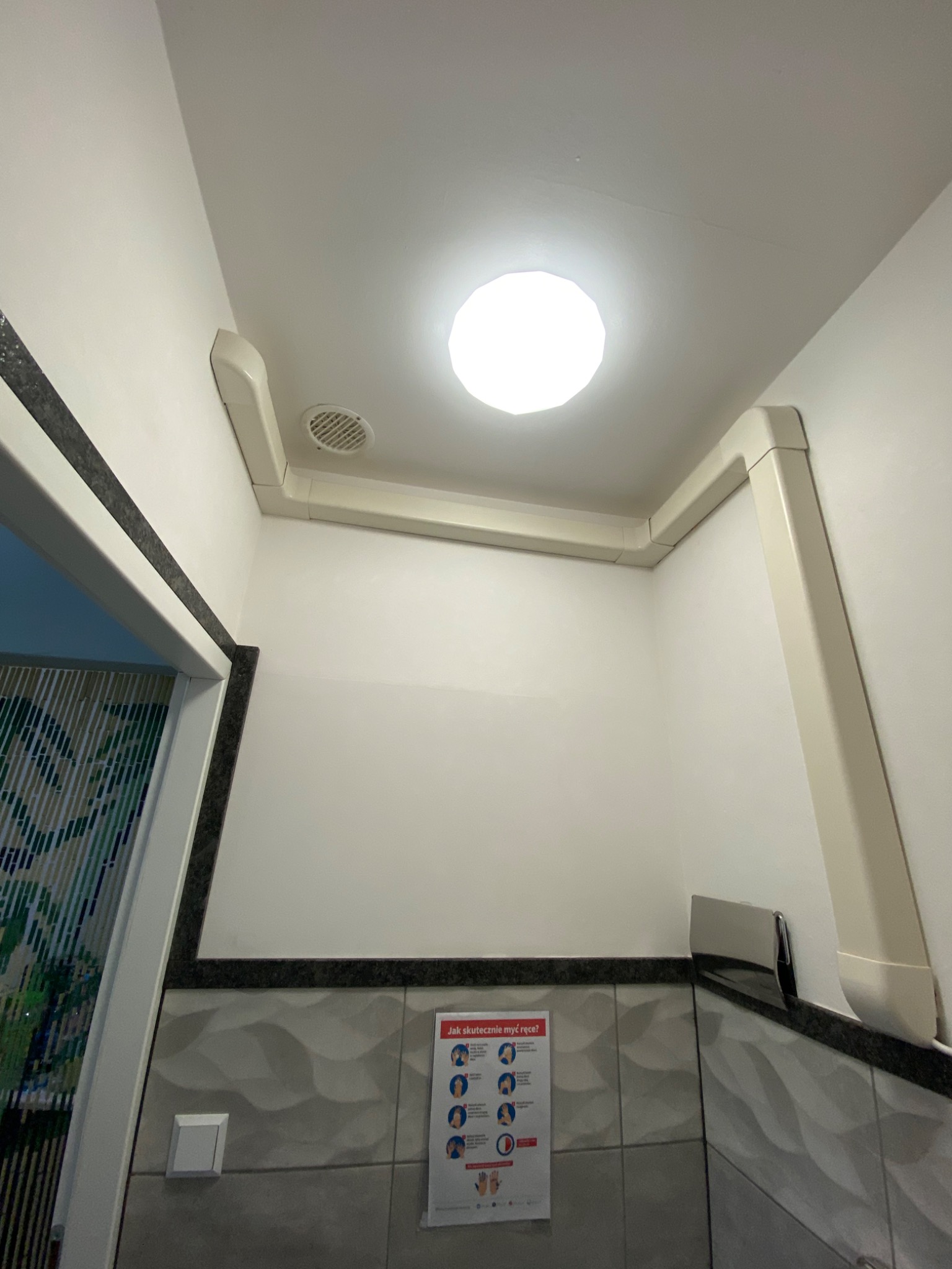 Białe kanały wentylacyjne poprowadzone pod sufitem w pomieszczeniu, widoczny także okrągły wlot wentylacyjny i plakat z instrukcją mycia rąk.