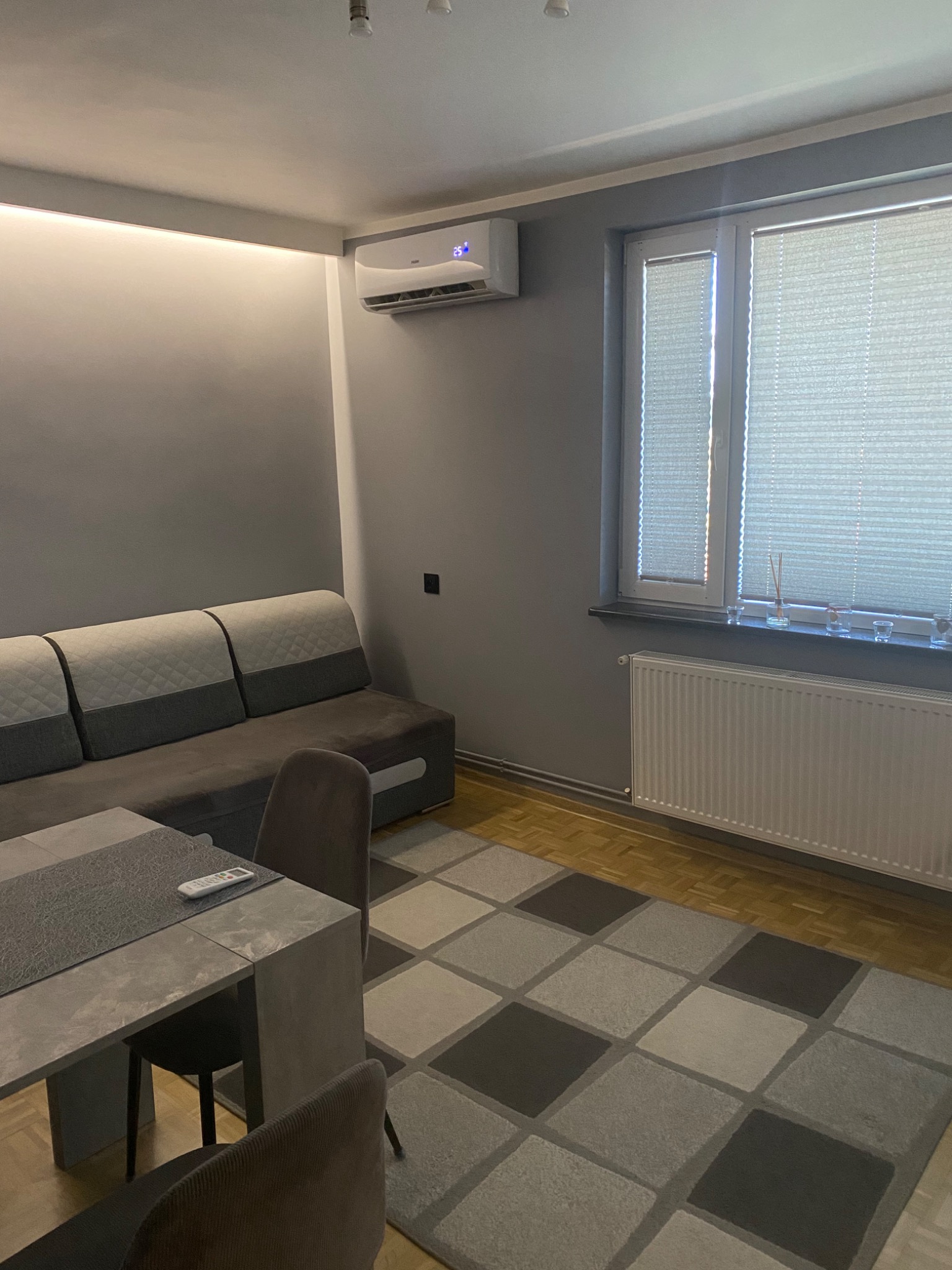 Wnętrze pokoju dziennego z zainstalowanym klimatyzatorem ściennym, widocznym nad sofą. Na stole leży pilot do klimatyzacji. Szare ściany i dywan w kratę.