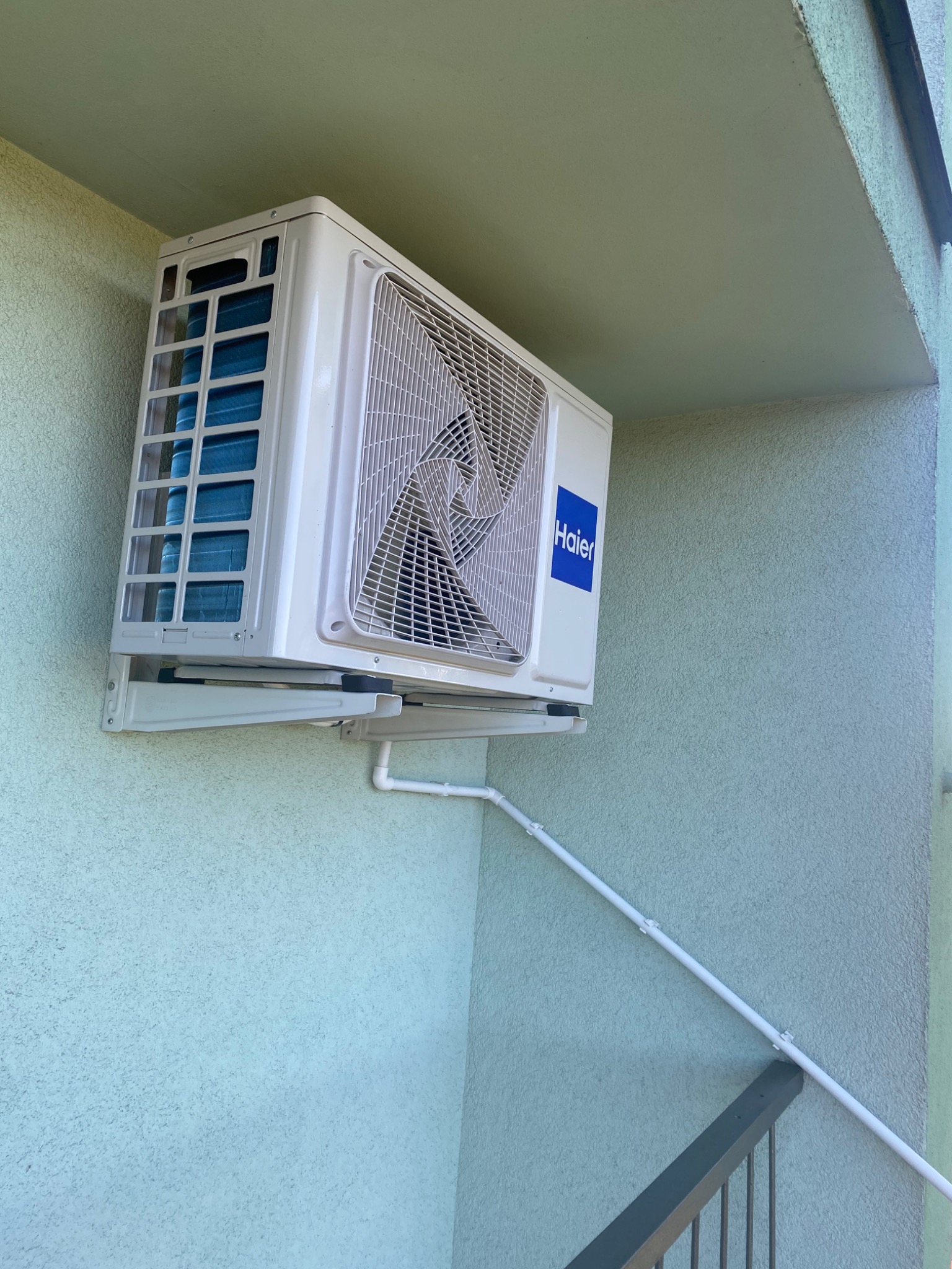 Zewnętrzna jednostka klimatyzacji Haier zamontowana na ścianie budynku o jasnej, zielonkawej elewacji, widoczne białe rurki odprowadzające skropliny.
