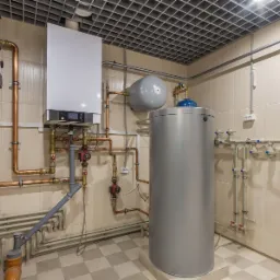 Kotłownia z nowoczesnym piecem gazowym, zbiornikiem buforowym i rozbudowaną instalacją hydrauliczną z miedzianych rur, widok na ścianę z beżowych płytek i kratownicowy sufit.