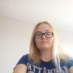 Portret kobiety z blond włosami i okularami w czarnych oprawkach, ubrana w niebieską koszulkę z nadrukiem, sfotografowana pod kątem od dołu.