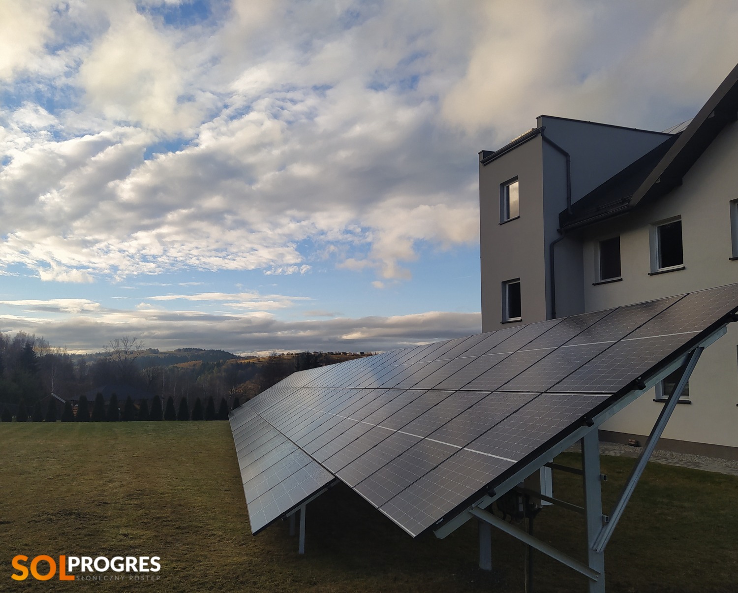 Panel słoneczny na trawniku przed nowoczesnym domem w słoneczny dzień z malowniczymi chmurami.