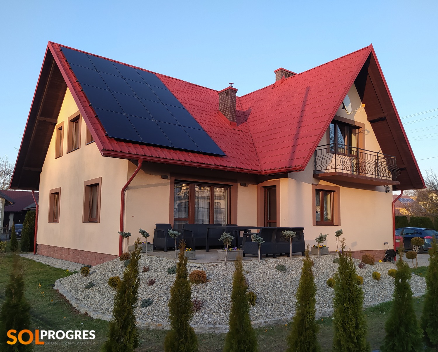 Dom jednorodzinny z czerwoną dachówką i panelami fotowoltaicznymi na dachu, widok z przodu, z zadbanym ogrodem.