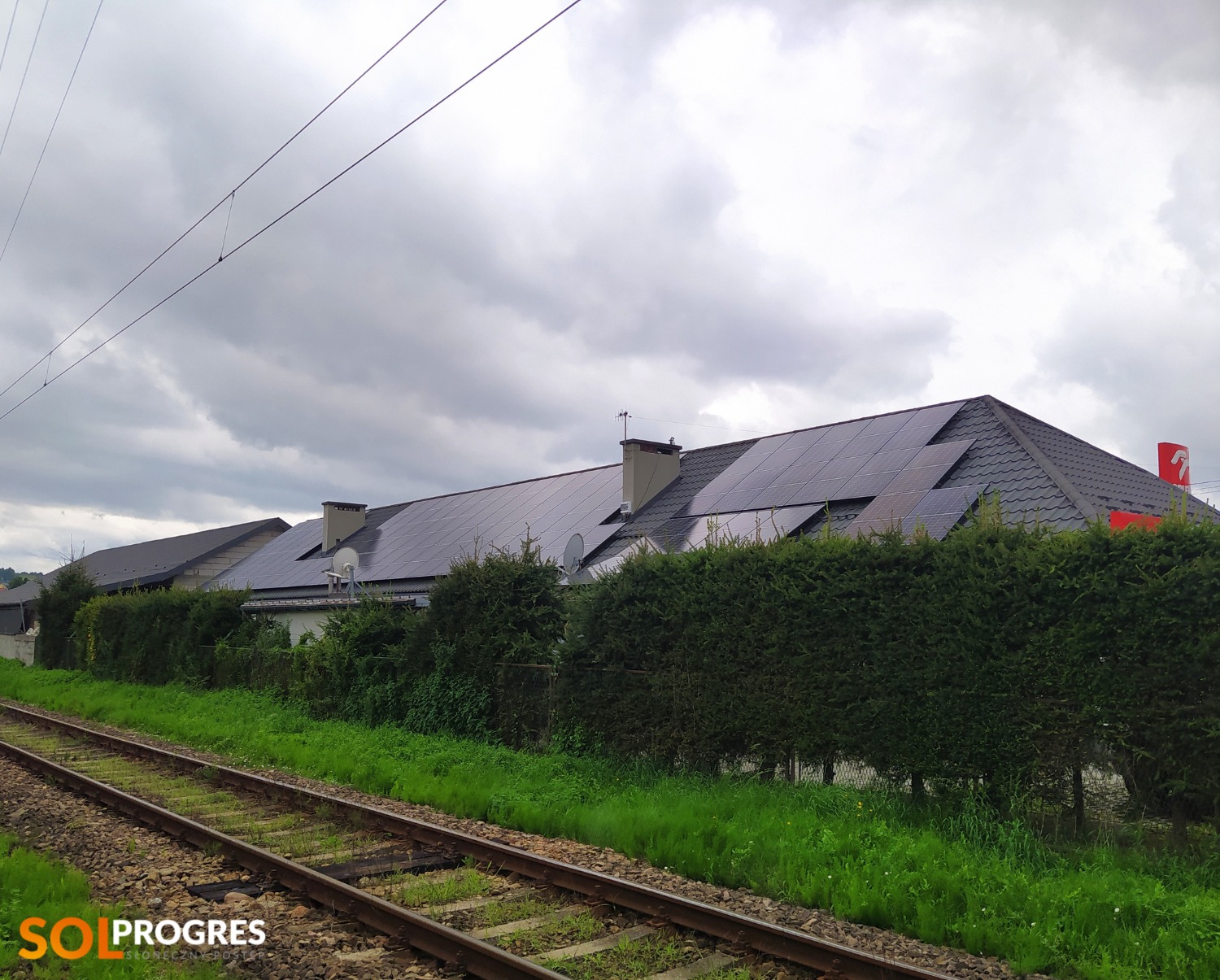 Długi budynek z panelami słonecznymi na dachu, widziany z perspektywy torów kolejowych, częściowo zasłonięty żywopłotem, pochmurne niebo.