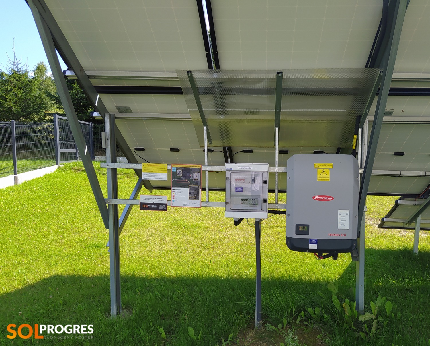 Widok od spodu na panele słoneczne z zamontowanym inwerterem Fronius ECO i skrzynką zabezpieczeń na tle trawnika i ogrodzenia.