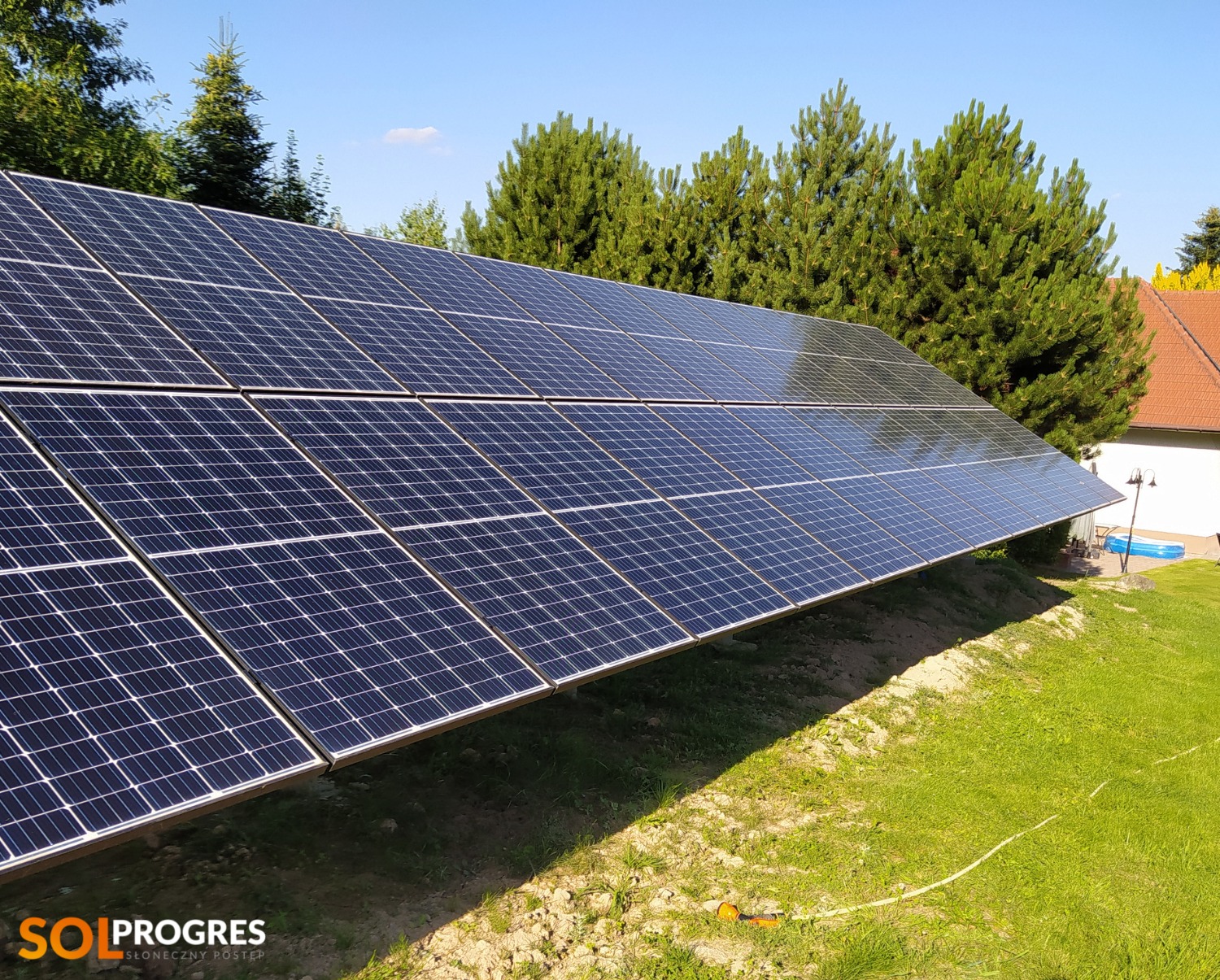 Instalacja paneli słonecznych na trawiastym terenie, w tle dom z czerwonym dachem i zielone drzewa iglaste, widoczne logo firmy SOL PROGRES.