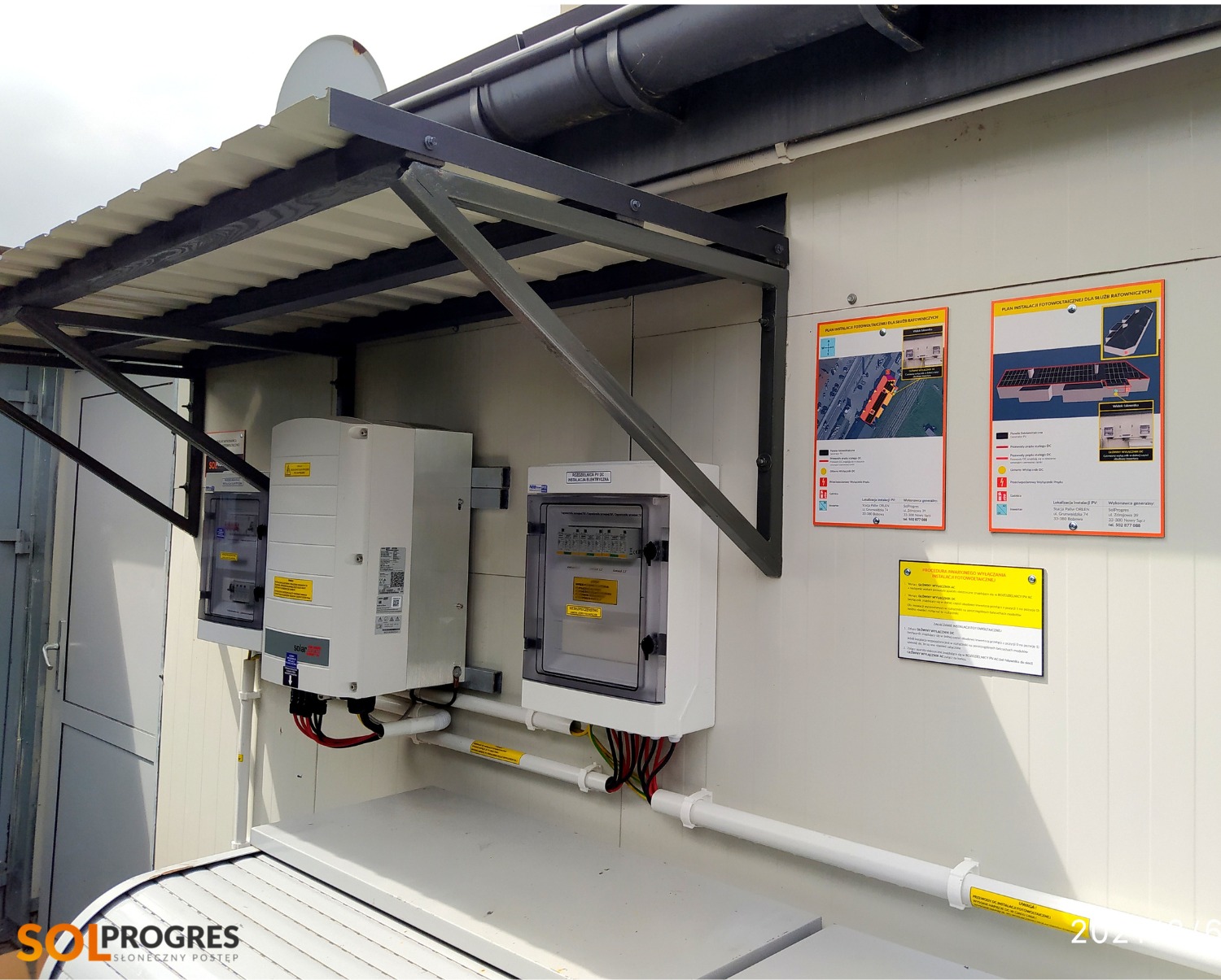 Zewnętrzna instalacja z dwoma białymi falownikami i skrzynką rozdzielczą pod zadaszeniem, widoczne schematy instalacji fotowoltaicznej na ścianie.