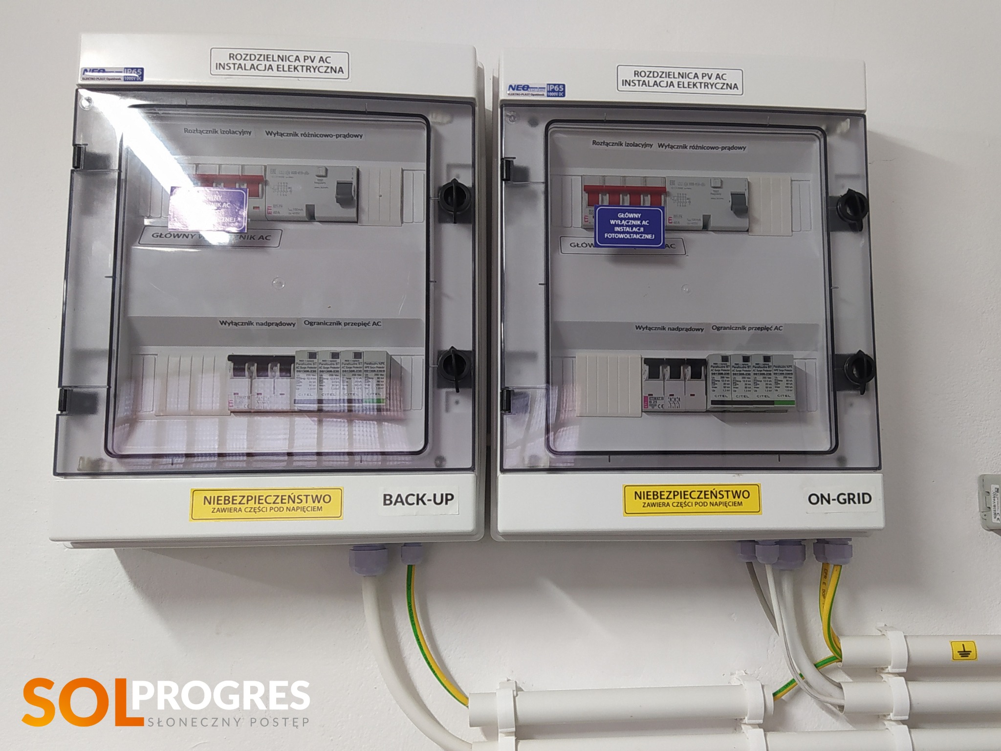 Dwie rozdzielnice PV AC, jedna oznaczona jako 'BACK-UP', druga 'ON-GRID', z widocznymi elementami instalacji elektrycznej i przewodami. Żółta naklejka ostrzegawcza informuje o niebezpieczeństwie...