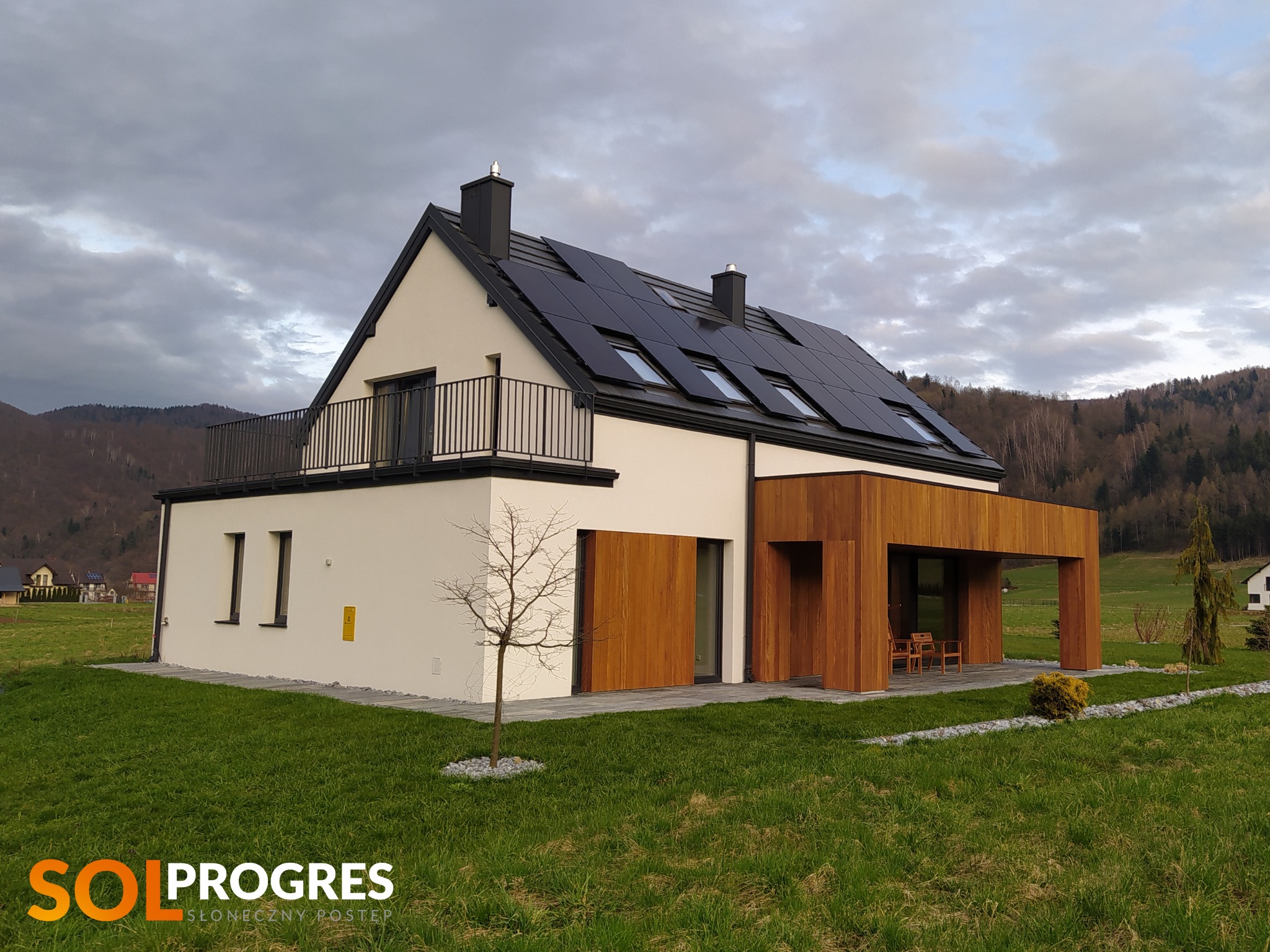 Nowoczesny dom jednorodzinny z panelami słonecznymi na dachu, balkonem z metalową balustradą i drewnianą elewacją, otoczony zielonym trawnikiem na tle gór i pochmurnego nieba. W rogu widoczne logo...