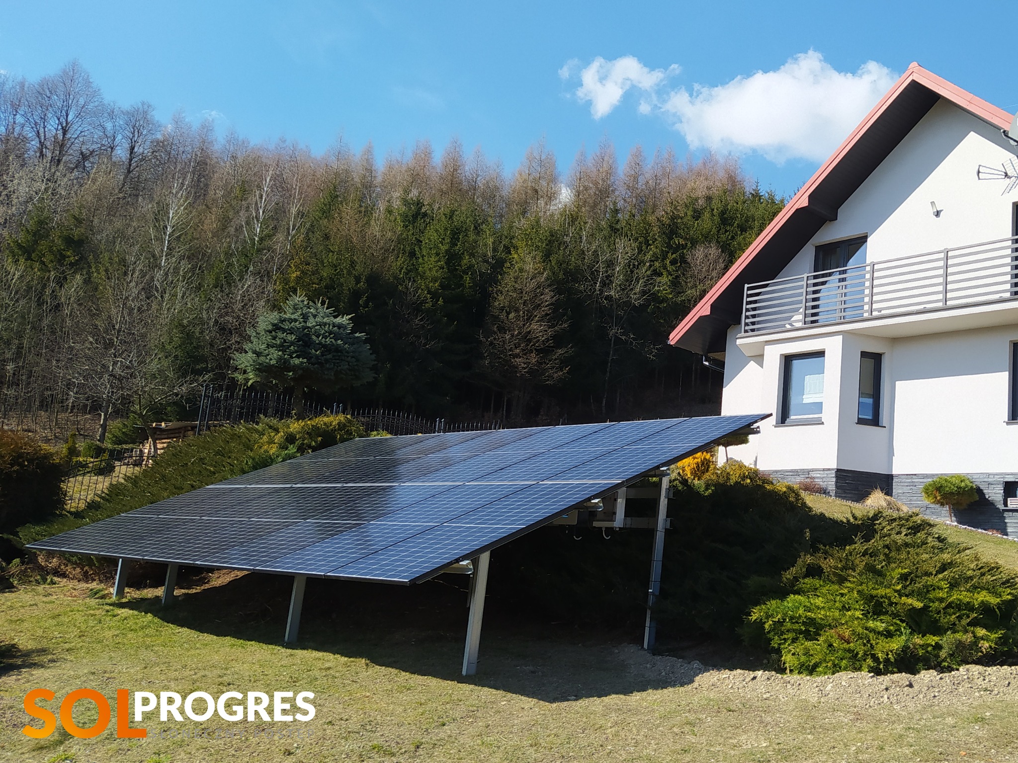 Panel słoneczny na gruncie przed domem jednorodzinnym z widocznym logo firmy instalacyjnej w rogu kadru.