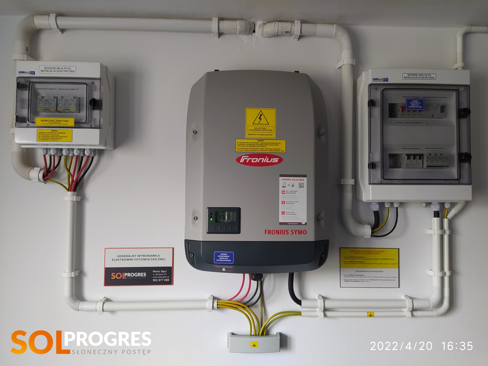 Instalacja fotowoltaiczna wewnątrz budynku: szary inwerter Fronius Symo, dwie rozdzielnice elektryczne PV DC i PV AC, białe rury ochronne na ścianie, tabliczka informacyjna firmy SolProgres.