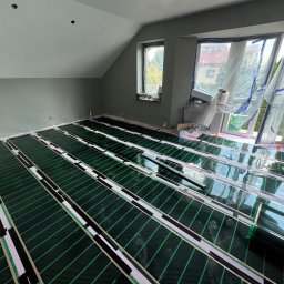 ElektroBud - Podłogówka elektryczna rozłożona w pokoju na poddaszu, widoczne folie grzewcze w kolorze zielonym, biała izolacja i czarne paski mocujące, okna zabezpieczone folią malarską, ściany w odcieniach...