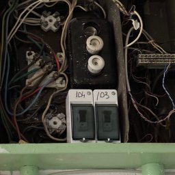 ElektroBud - Otwarta skrzynka elektryczna z bezpiecznikami topikowymi i dwoma wyłącznikami nadprądowymi oznaczonymi numerami 104 i 103, widoczne liczne kable w różnych kolorach i złączki.