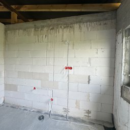ElektroBud - Surowy stan wnętrza w trakcie prac: ściany z białych bloczków, widoczne przewody elektryczne z czerwonymi puszkami podtynkowymi, instalacja podłogowa w peszlach, betonowa wylewka, drewniana...