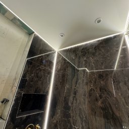 ElektroBud - Łazienka w trakcie wykańczania: ciemny marmur z podświetleniem LED, złota bateria podtynkowa, wnęka w ścianie, sufit z oświetleniem punktowym.