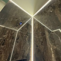 ElektroBud - Ściany i sufit wykończone marmurem z wbudowanym oświetleniem LED tworzącym geometryczny wzór. Widoczna puszka elektryczna w ścianie.