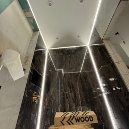 ElektroBud - Łazienka w trakcie wykończenia, widoczne marmurowe płytki, oświetlenie LED w kształcie trójkątów, złota armatura, kartony z napisem WOOD i fragmenty instalacji elektrycznej na ścianie.