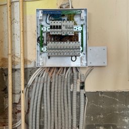 ElektroBud - Otwarta skrzynka bezpiecznikowa z licznymi przewodami w peszlach, zamontowana na częściowo otynkowanej ścianie w trakcie prac remontowych.