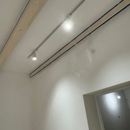 ElektroBud - Biały sufit z zamontowanymi drewnianymi belkami i szynoprzewodem z reflektorami punktowymi, widoczne wiszące przewody elektryczne oraz fragment ościeżnicy drzwiowej.