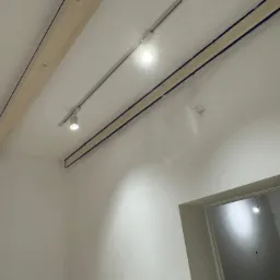 Biały sufit z zamontowanymi drewnianymi belkami i szynoprzewodem z reflektorami punktowymi, widoczne wiszące przewody elektryczne oraz fragment ościeżnicy drzwiowej.