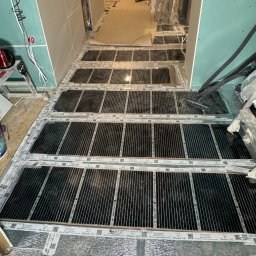 ElektroBud - Montaż folii grzewczej pod podłogą w trakcie wykańczania wnętrza, widoczne panele folii grzewczej z etykietami 'Mission Air' rozłożone na folii aluminiowej, w tle ściany z płyt gipsowo-kartonowych...