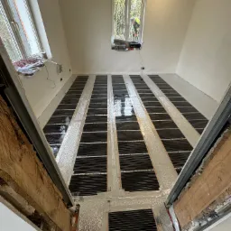 Rozłożone maty grzewcze na folii aluminiowej w pomieszczeniu w trakcie prac wykończeniowych, widok z perspektywy progu drzwiowego, okno z widokiem na zewnątrz w tle.