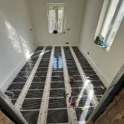 Montaż ogrzewania podłogowego elektrycznego w nowym, jasnym pomieszczeniu z dwoma oknami dachowymi, widoczne narzędzia i materiały instalacyjne.