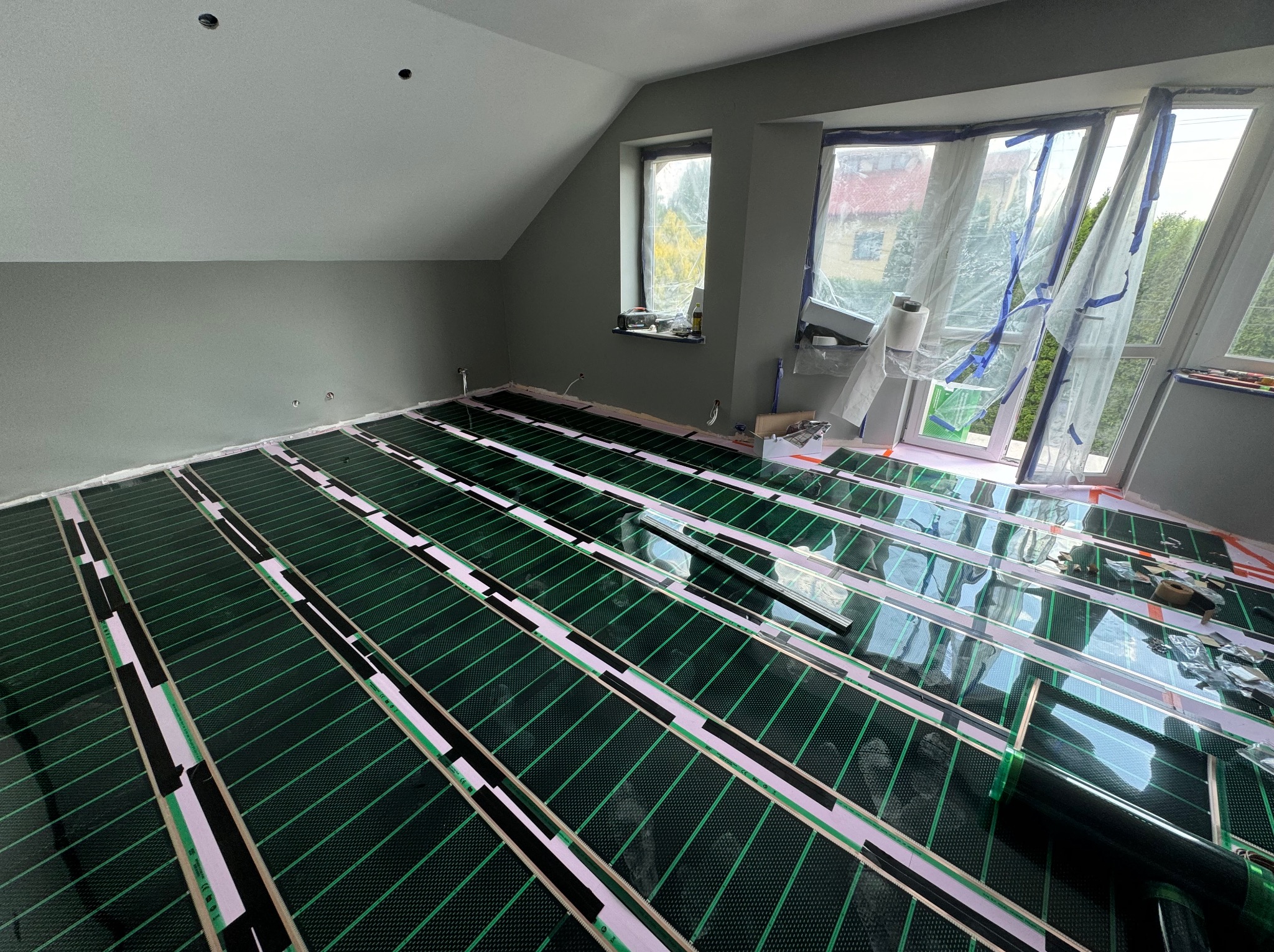 Podłogówka elektryczna rozłożona w pokoju na poddaszu, widoczne folie grzewcze w kolorze zielonym, biała izolacja i czarne paski mocujące, okna zabezpieczone folią malarską, ściany w odcieniach...