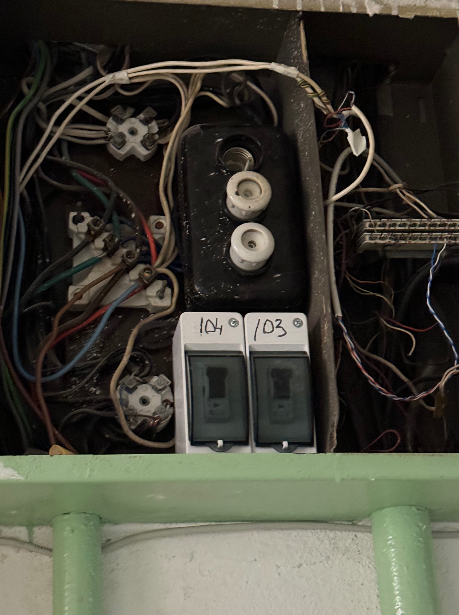 Otwarta skrzynka elektryczna z bezpiecznikami topikowymi i dwoma wyłącznikami nadprądowymi oznaczonymi numerami 104 i 103, widoczne liczne kable w różnych kolorach i złączki.